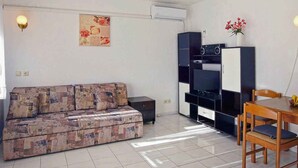 A living room with a sofa, a dining table with chairs, a TV, and a kitchen area.