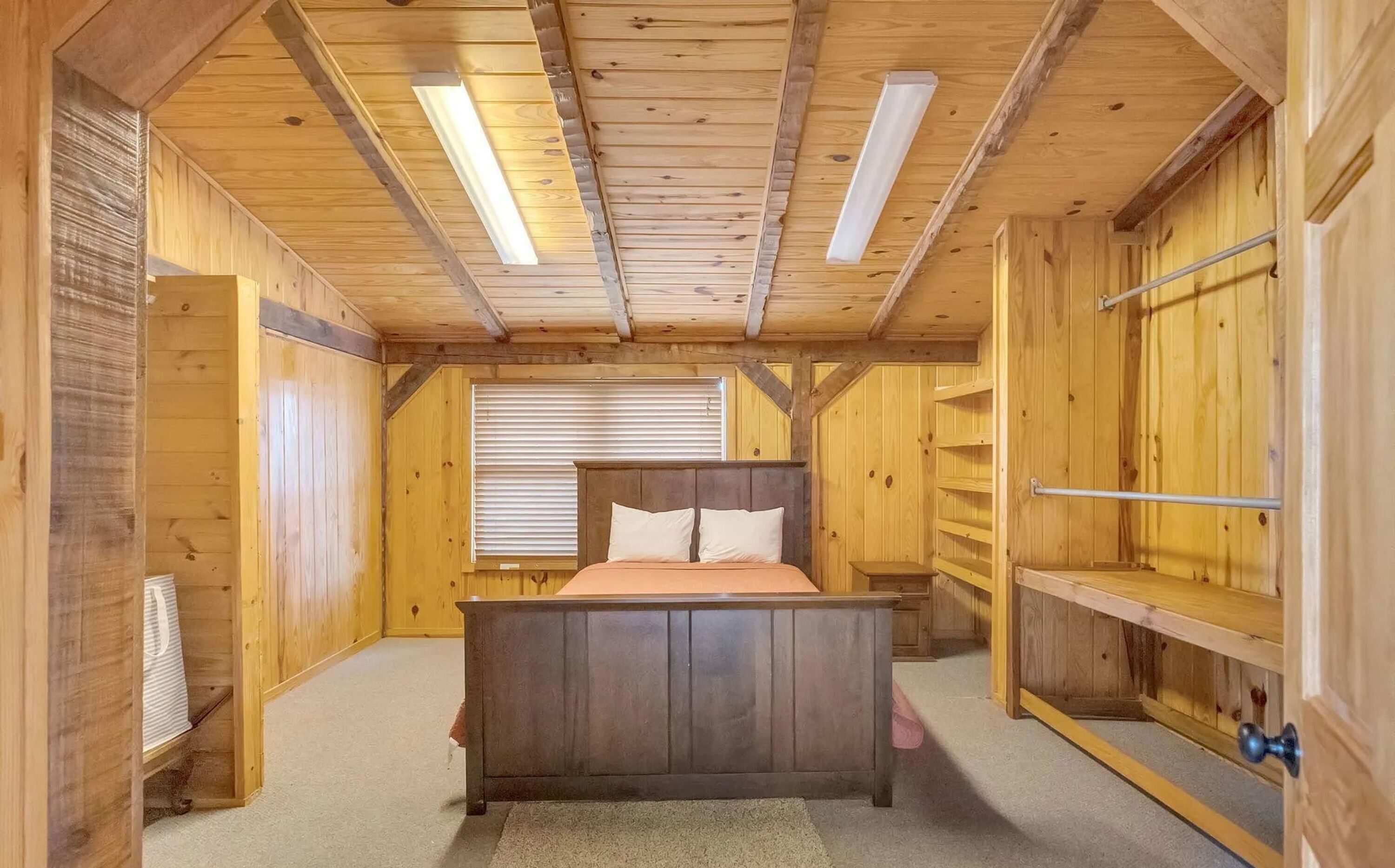 A wooden bedroom with a bed, a window with blinds, and built-in shelving.