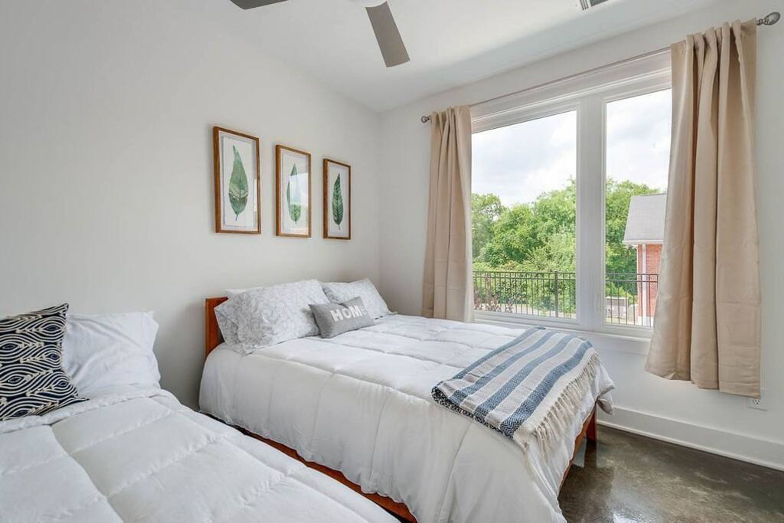 A bedroom with two beds, a large window with curtains, and framed botanical prints on the wall.