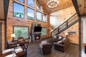 A spacious living room with a fireplace, wooden walls, a staircase, and large windows. - Luxury Brand New Cabin 5b 5 5ba w Arcade Theater (Sevierville)