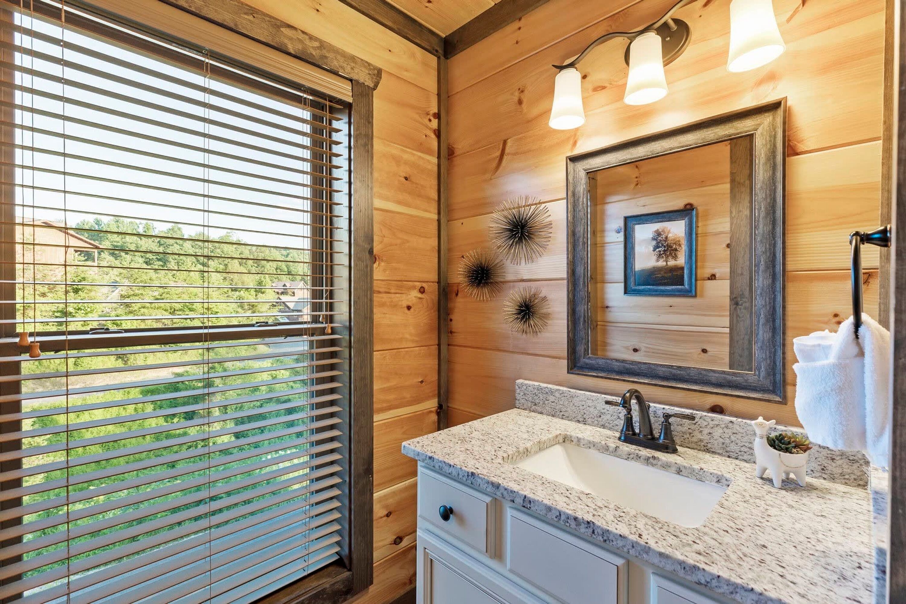 Un baño con paredes de madera, una ventana con persianas, un lavamanos con encimera de granito y un espejo con detalles decorativos.