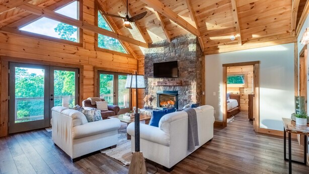 A spacious living room with a stone fireplace, wooden beams, and large windows. - Luxury 3 2 5 Cabin Lake Views Theatre Gameroom (Bryson City)