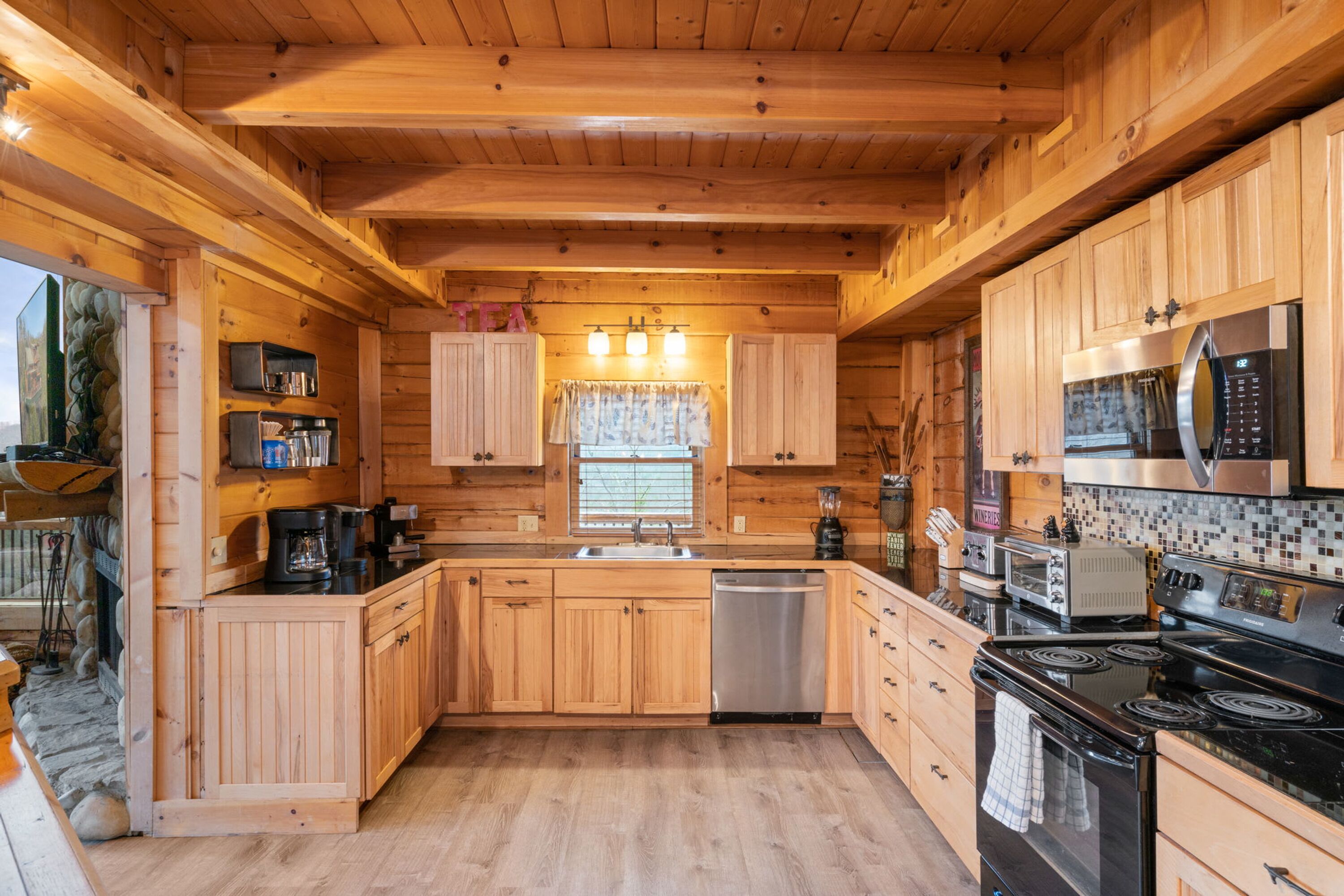 A wooden cabin kitchen with a dishwasher, stove, microwave, and a window with curtains.