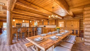 Ein Interieur einer Blockhütte mit einem langen Esstisch, gedeckt für sechs Personen, Holzbarhocker, einer Wand aus Naturstein und einer Küchenzeile mit modernen Geräten.