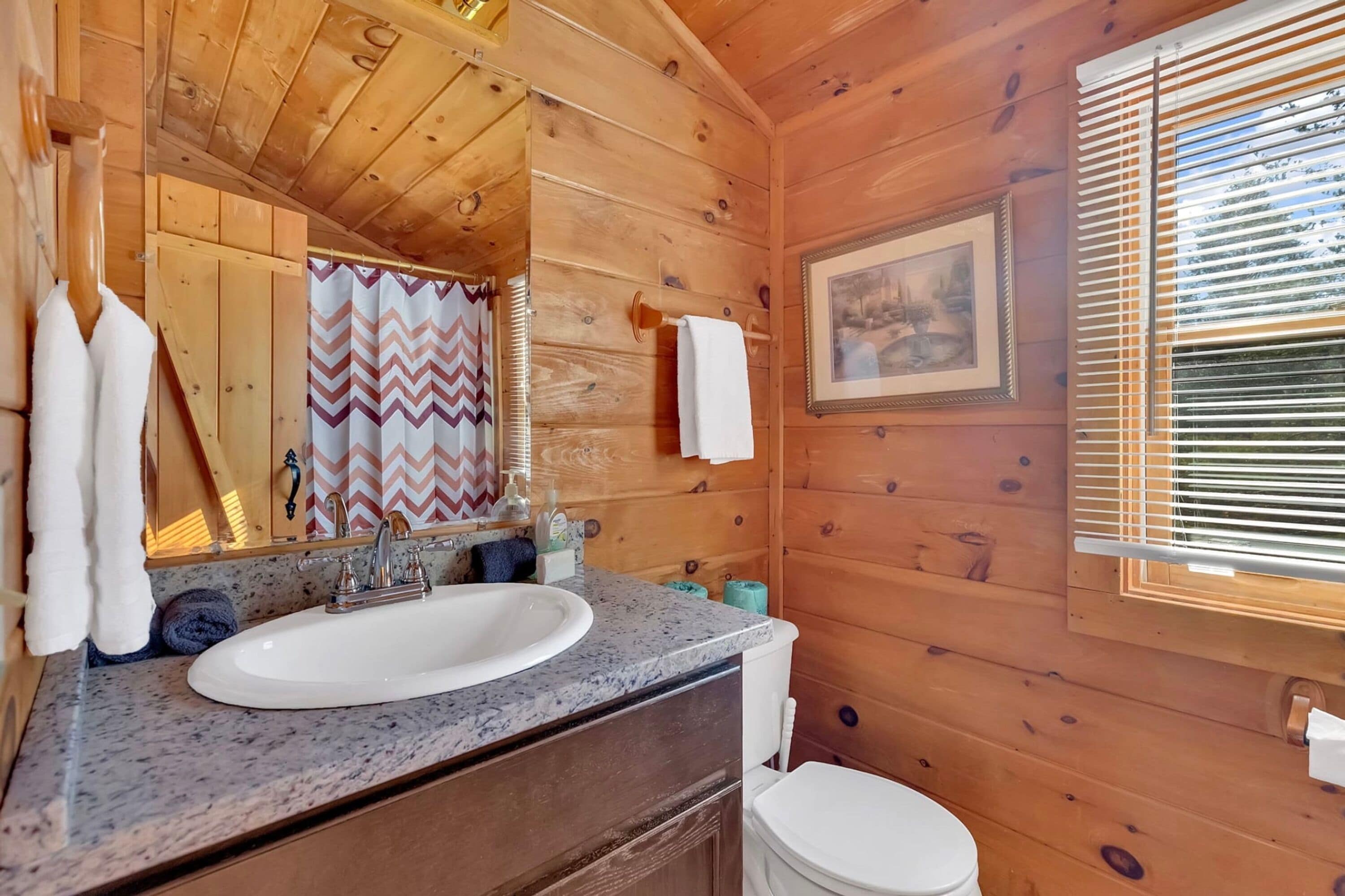 A wooden cabin bathroom with a granite sink, a mirror, a framed picture, and a window with blinds.