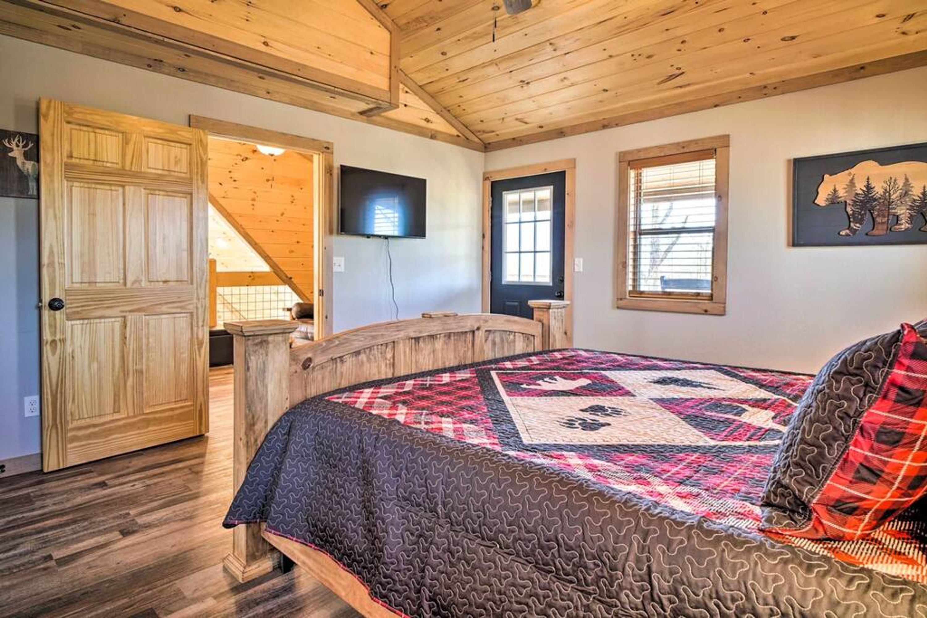 A bedroom with a wooden ceiling, a bed with a patterned quilt, a television on the wall, and a door leading to another room.
