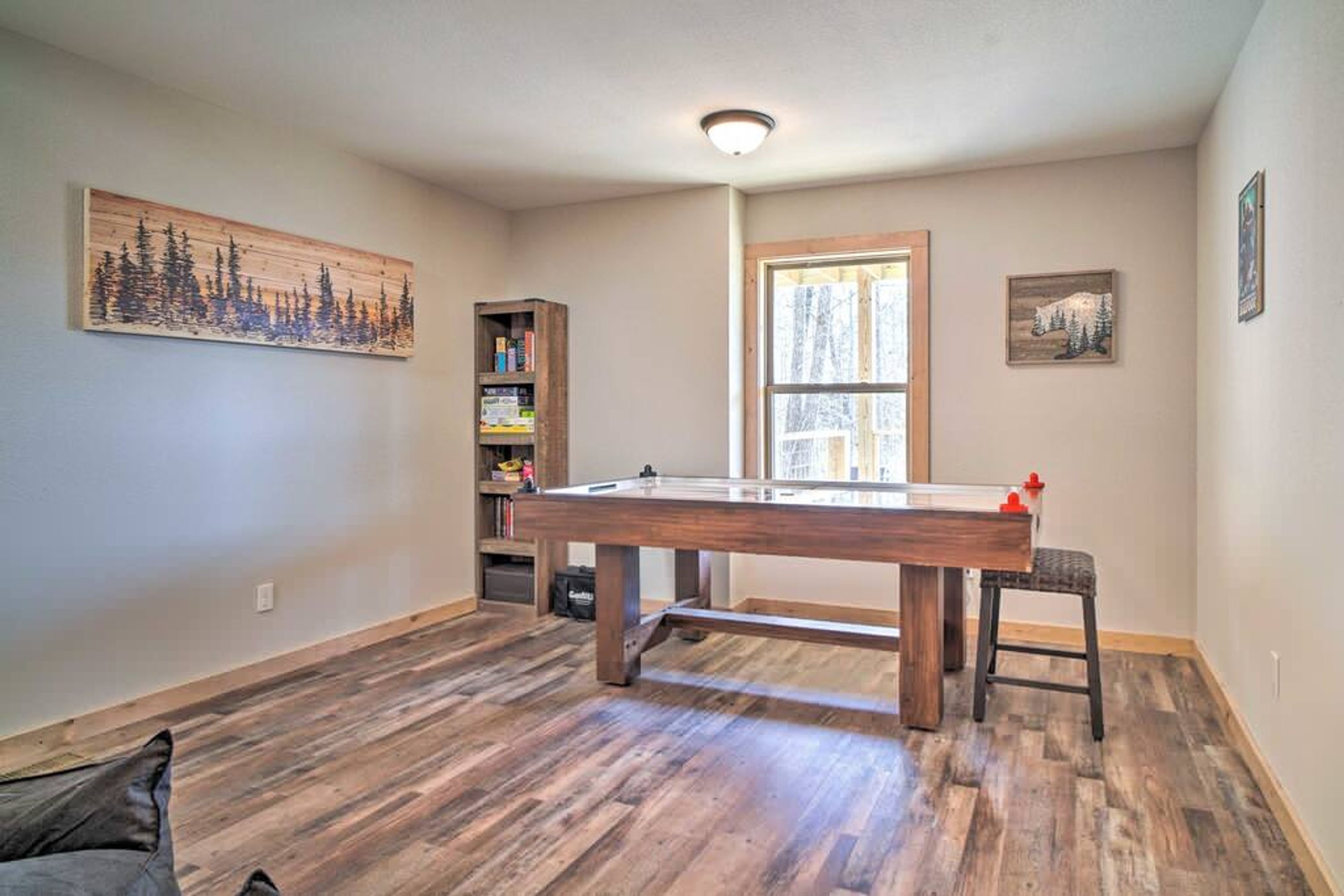 A room with a pool table, wooden flooring, a bookshelf, and a window with a view.