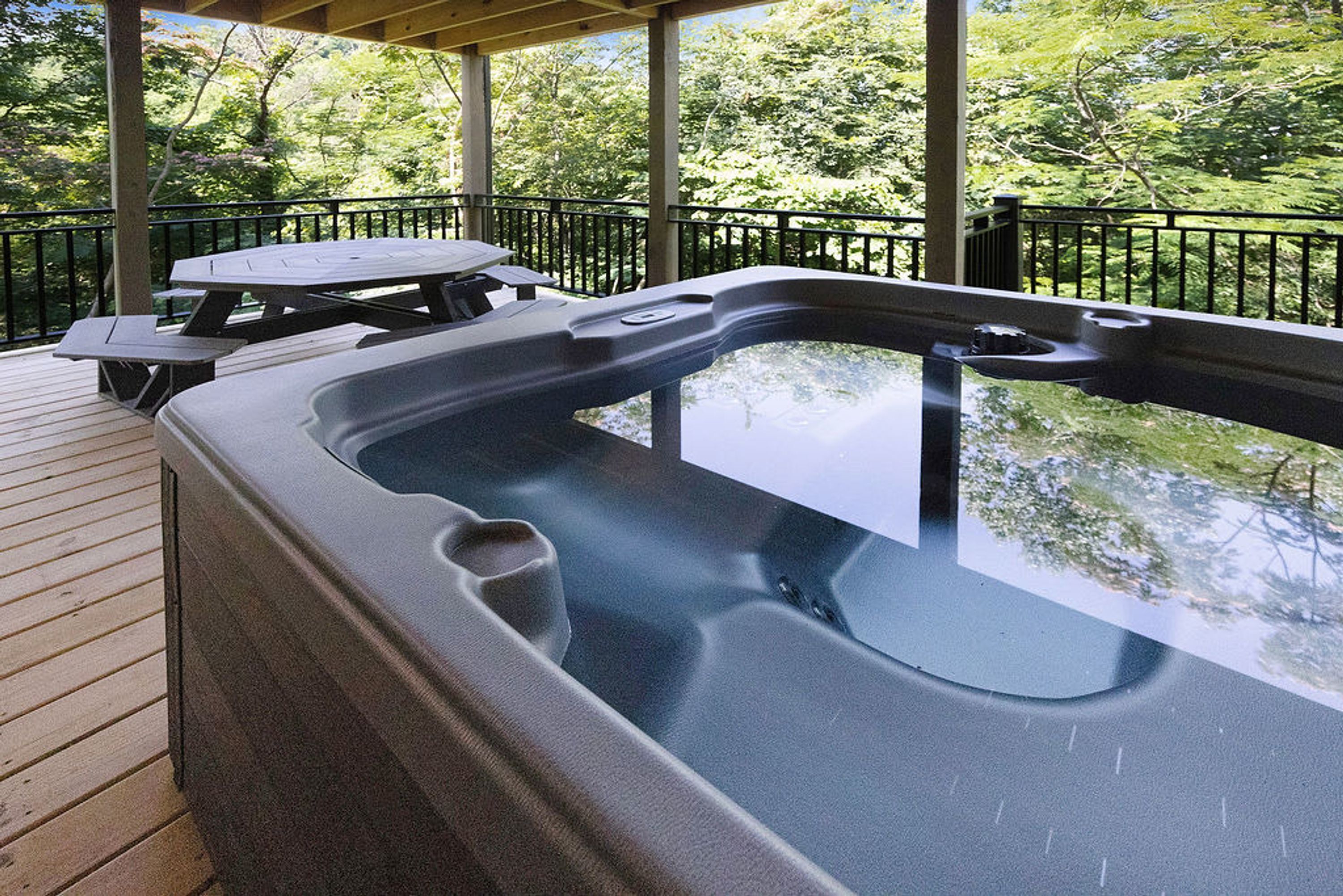 A hot tub on a wooden deck with a picnic table nearby, surrounded by a metal railing and trees.