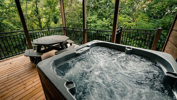 A hot tub on a wooden deck with a table and chairs, surrounded by a railing and trees.
