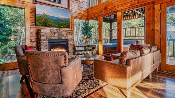 A cozy cabin interior with a stone fireplace, plush seating, and large windows offering scenic views. - Mountain Majesty With Spectacular Views (Sevierville)