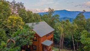 A two-story log cabin with a balcony, surrounded by dense forest and mountains in the background. - Mountain Majesty With Spectacular Views (Sevierville)
