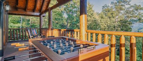 A wooden deck with a foosball table and a pool table, surrounded by a wooden railing and overlooking a forested area.