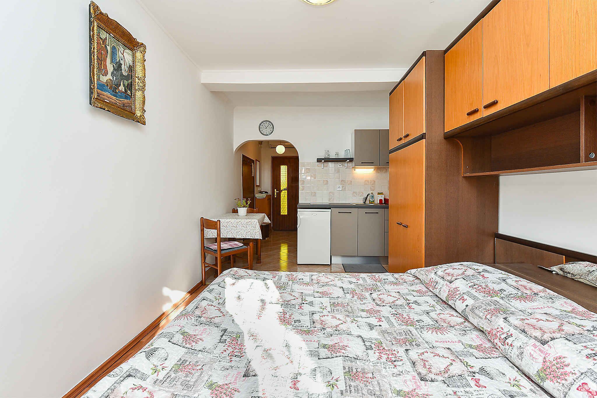 A compact bedroom with a bed, wooden wardrobe, and a small kitchenette in the background.