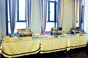 Une table de buffet garnie de divers plats et accompagnée du matériel de service nécessaire, placée devant une fenêtre ornée de rideaux.