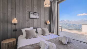 A bedroom with a large bed, two wall-mounted lamps, a framed picture, and a view of mountains through a window.