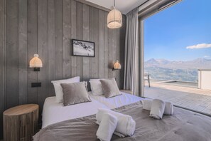 A bedroom with a large bed, two wall-mounted lamps, a framed picture, and a view of mountains through a window. - TERRESENS - Le Quartz (Peisey-Nancroix)