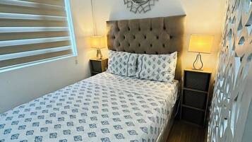 A bedroom with a tufted headboard, a patterned bedspread, two bedside lamps, and a decorative mirror on the wall.