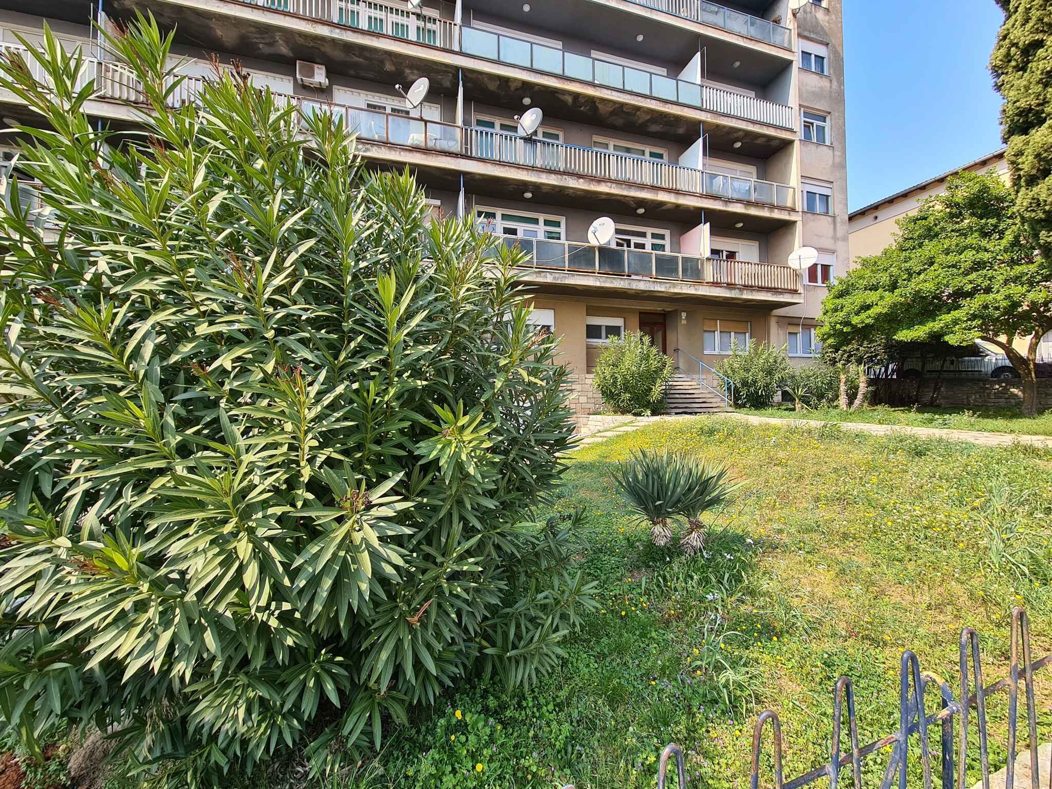 A multi-story residential building with balconies, satellite dishes, and a well-maintained garden area with various plants and flowers.