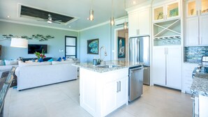 A modern kitchen with white cabinetry, a central island with a sink, and a dining area with a sofa and TV. - Beachfront Condo Complex In Placencia Village (Placencia)