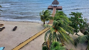 Una playa con un muelle de madera que lleva a una estructura con techo de paja, palmeras y una vista despejada del océano.