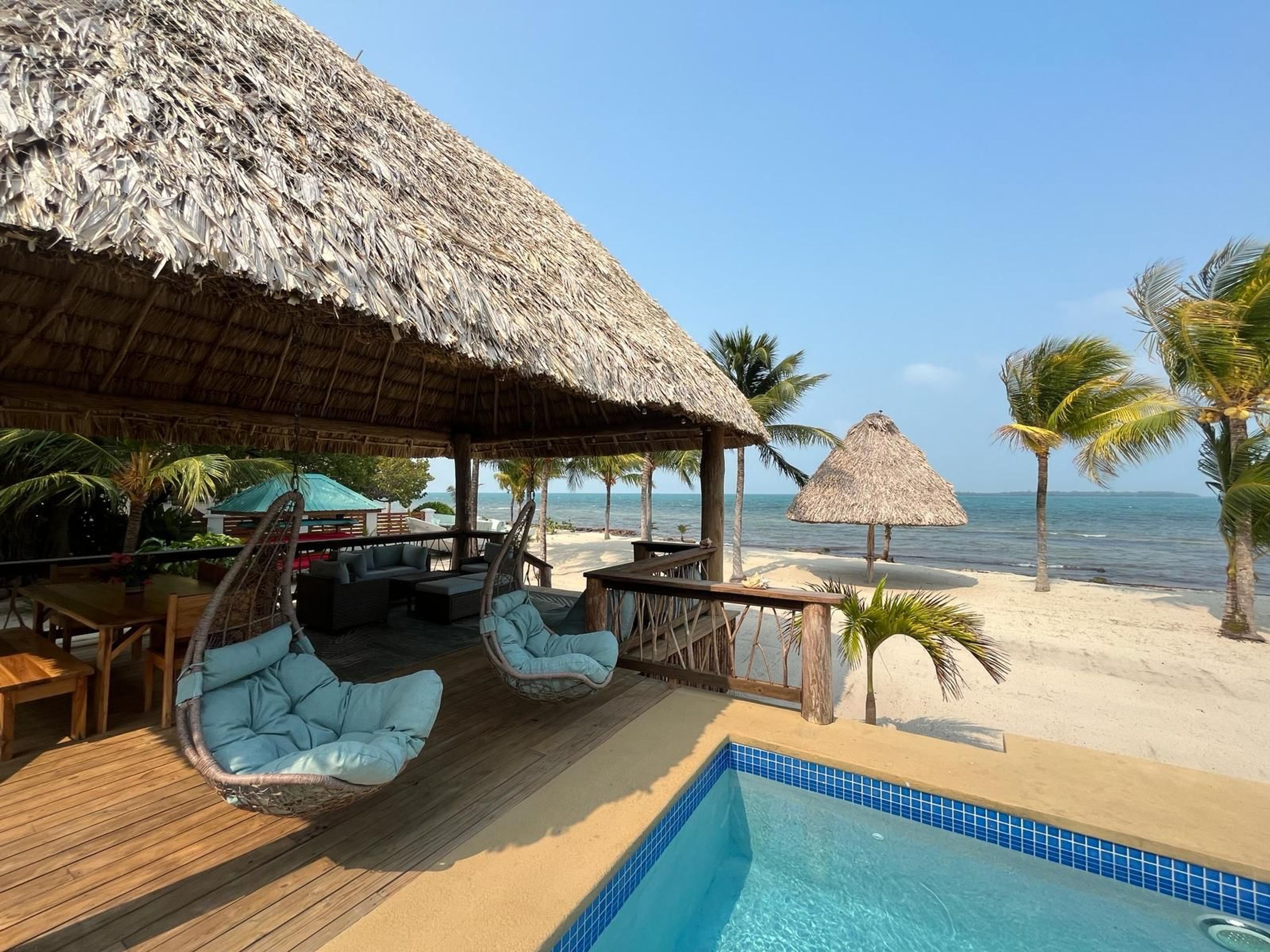 A beachside lounge with a pool, thatched-roof structures, and palm trees.