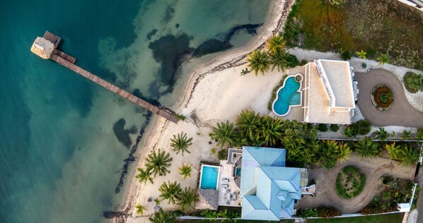 An aerial view of a coastal property with a beach, a swimming pool, and a house with a blue roof.