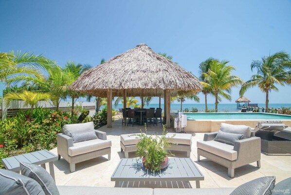 A beachside lounge with a thatched umbrella, outdoor seating, and a pool. - Luxury Oceanfront Villa w 2 Pools Beach Pier (Placencia)