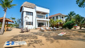 A modern two-story building with a thatched roof, surrounded by palm trees and a sandy beach area with lounge chairs.