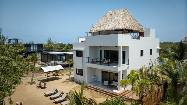 A modern two-story house with a thatched roof, multiple balconies, and a pool area with sun loungers.