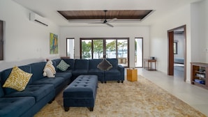 A spacious living room with a dark blue sectional sofa, a matching ottoman, and a patterned rug. There is a ceiling fan, a wooden floor, and a view of the outdoors through large windows.