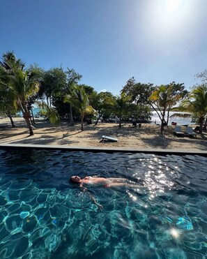 Piscina cristalina con una persona nadando, rodeada de palmeras y una zona de playa.