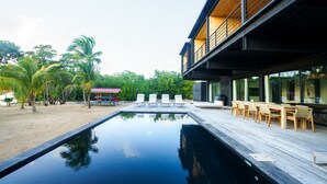 A modern pool area with a wooden deck, lounge chairs, and a view of a beach and palm trees. - NEW Modern Ocean Front Villa on Private Island (Placencia)