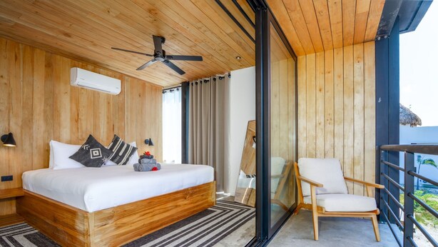 Une chambre avec un lit en bois, un ventilateur de plafond, une grande fenêtre avec des rideaux, un balcon avec une chaise et une vue sur l’océan.