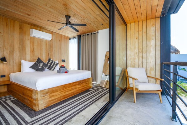 Une chambre avec un lit en bois, un ventilateur de plafond, une grande fenêtre avec des rideaux, un balcon avec une chaise et une vue sur l’océan.