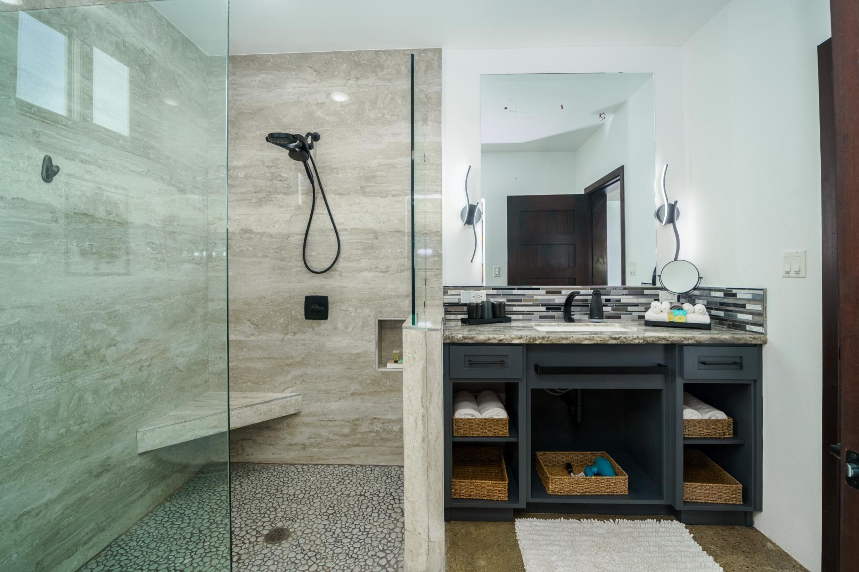 A modern bathroom with a glass shower enclosure, a marble-tiled wall, a dark vanity with wicker baskets, and a large mirror.