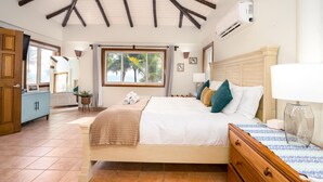 A bedroom with a bed, bedside table, lamp, and a window with a view of palm trees.