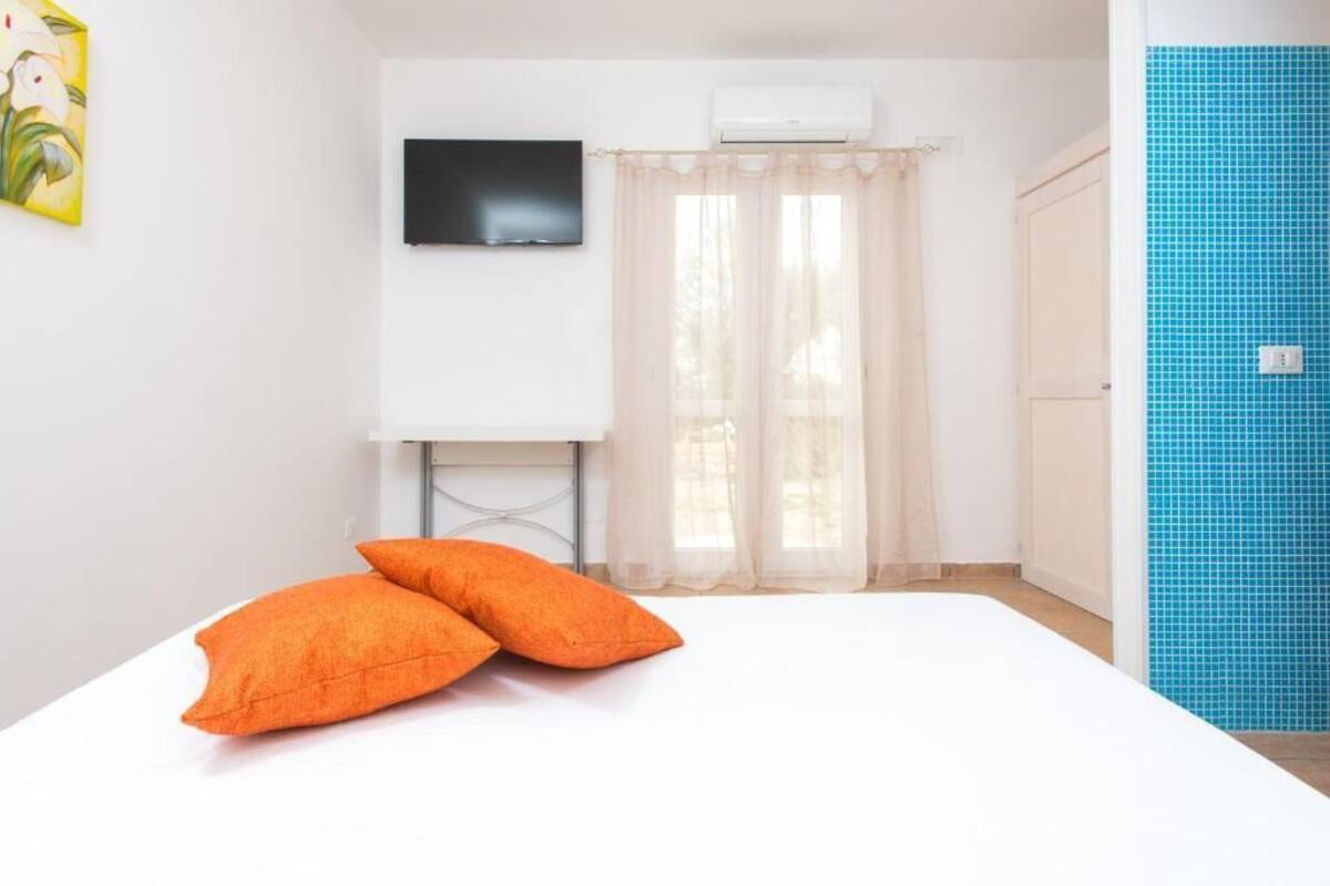 A room with a bed, two orange pillows, a flat-screen TV on the wall, a window with sheer curtains, and a blue tiled wall.