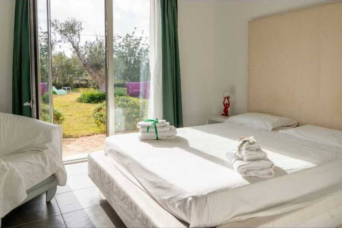 A hotel room with a large bed, a bedside table, and a view of a garden through a sliding glass door.