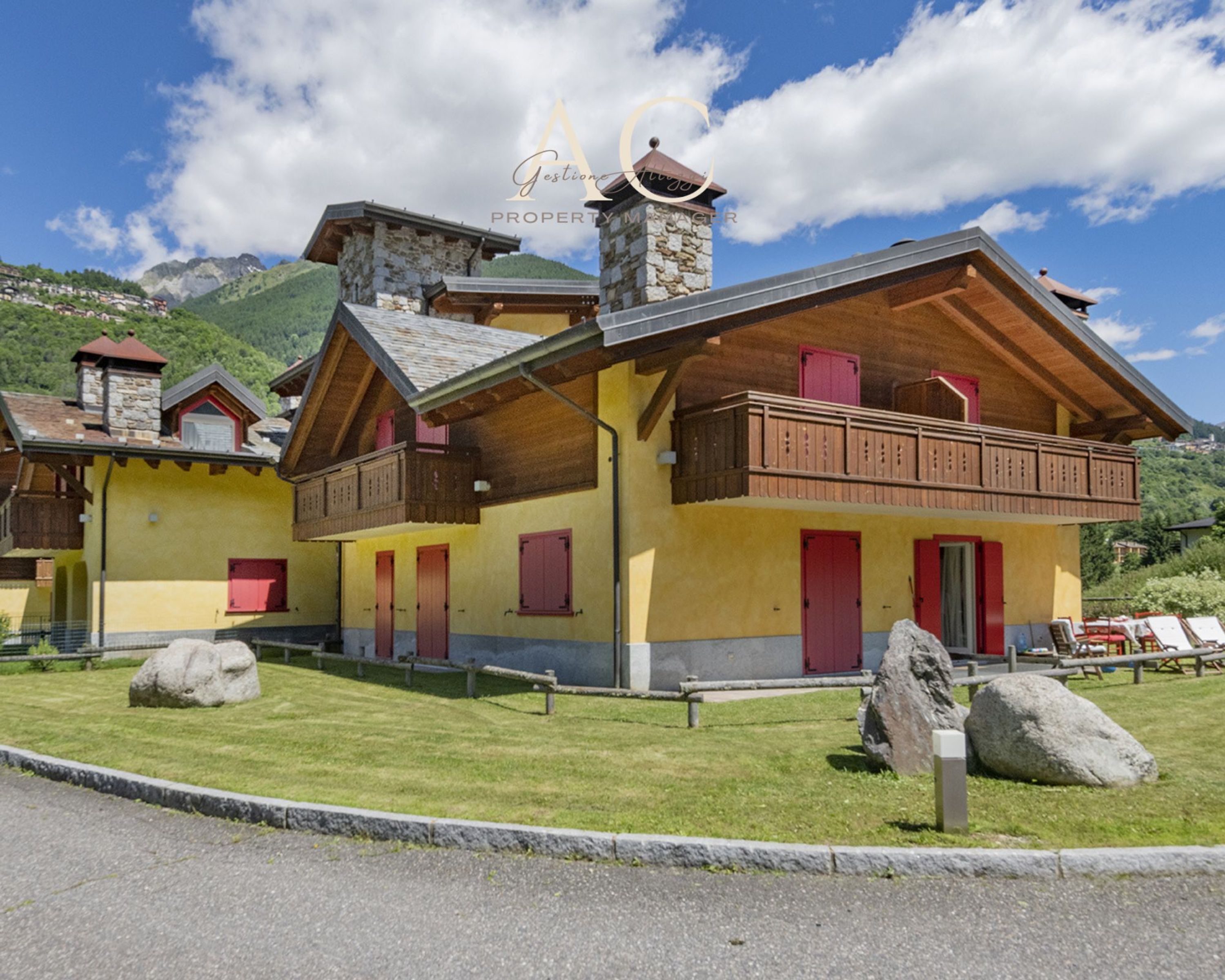 Un edificio de estilo chalé con balcón de madera, chimenea de piedra y montañas al fondo.