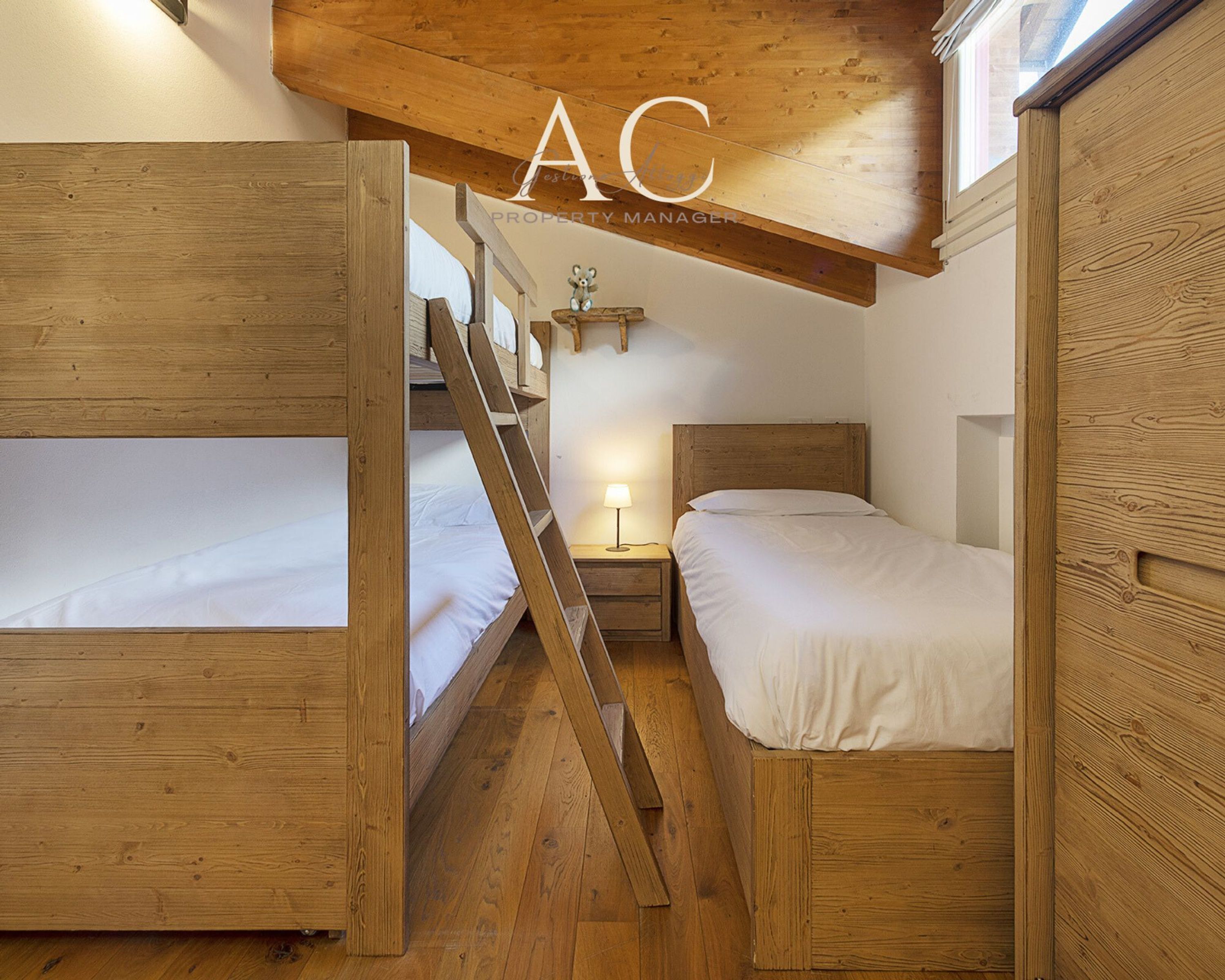 Habitación con literas de madera, una escalera, una mesita de noche con lámpara y un cabecero de madera.