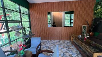 A room with a wooden panel wall, a tiled floor, a wooden table with a vase of flowers, and a large window with a view of greenery outside.