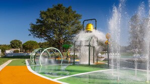 Una fontana d'acqua con getti multipli, circondata da un'area giochi verde e un sentiero giallo.