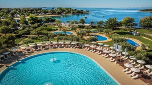 Un resort con una grande piscina centrale circondata da lettini e ombrelloni, un porto turistico con barche e una vegetazione lussureggiante.
