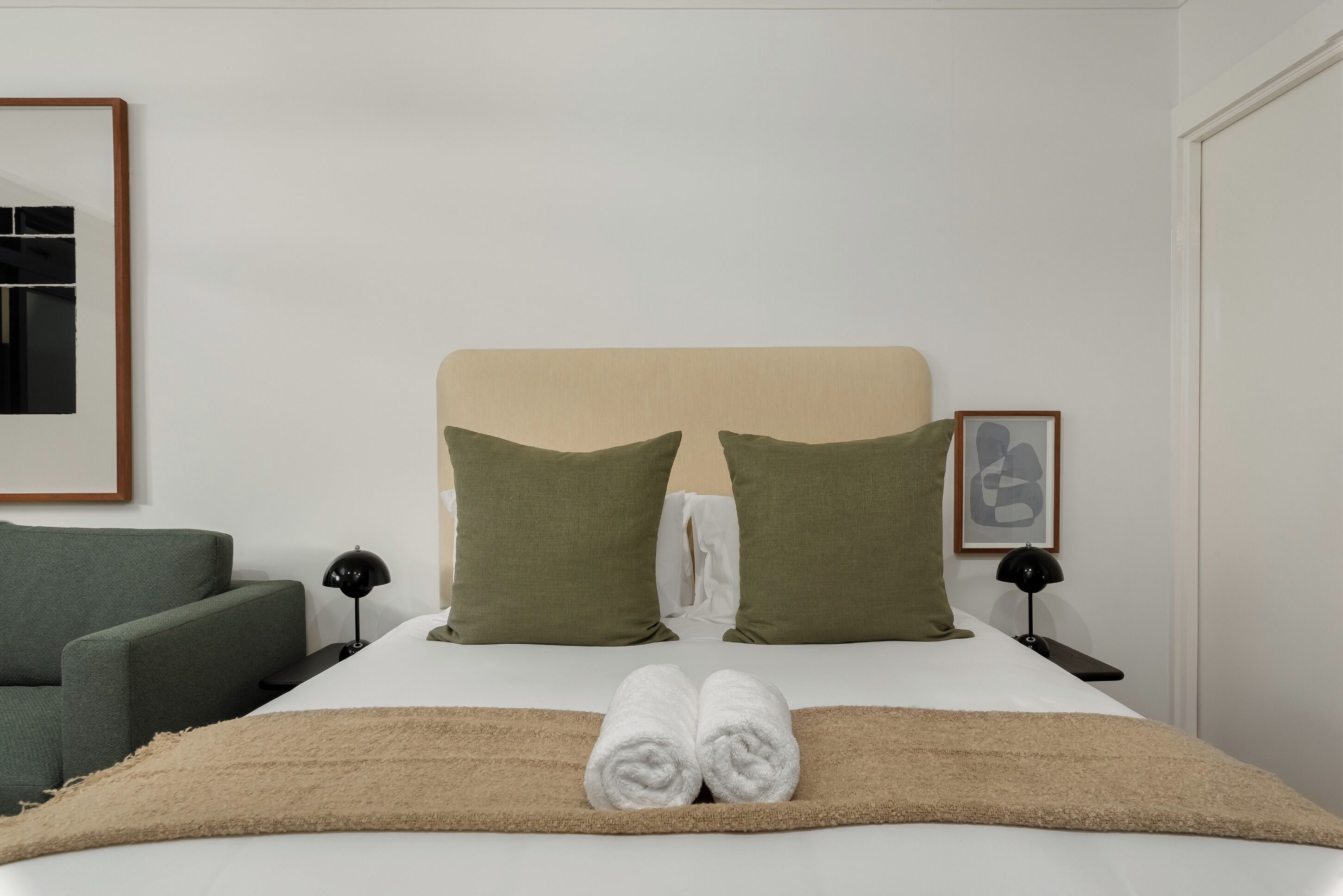 A neatly made bed with two pillows, a pair of towels, a bedside lamp, and a framed picture on the wall.