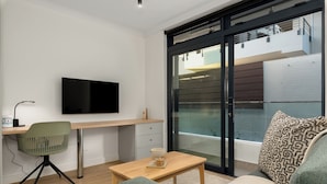 A modern living room with a flat-screen TV, a wooden desk, a green chair, a sofa with patterned cushions, and a sliding glass door leading to an outdoor area. - The Sage | Studio With Courtyard Access (Cape Town)