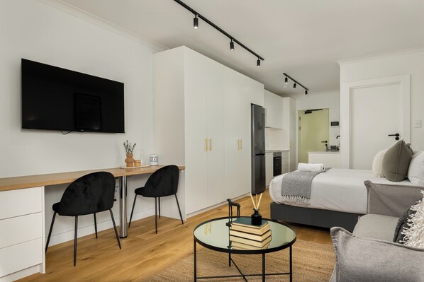 Una habitación moderna y minimalista con televisión de pantalla plana montada en la pared, un escritorio de madera con dos sillas negras, una mesa de cristal con libros y una vela, una cama con cabecero gris, y una pequeña cocina americana al fondo.