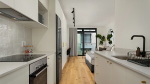 Cozinha moderna com armários brancos, fogão preto e piso de madeira. Há uma área de refeições com mesa e cadeiras, e uma cama com cabeceira branca ao fundo.