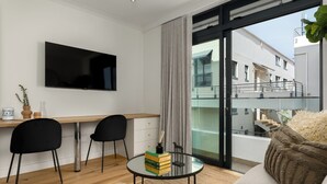 Uma sala de estar moderna com uma televisão de tela plana, uma escrivaninha de madeira, duas cadeiras pretas, uma mesa de centro de vidro com livros, um sofá bege e uma varanda com vista para edifícios.