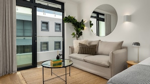 Sala de estar moderna com sofá, espelho redondo, mesa pequena com livros, planta e vista de um edifício por uma porta de vidro.