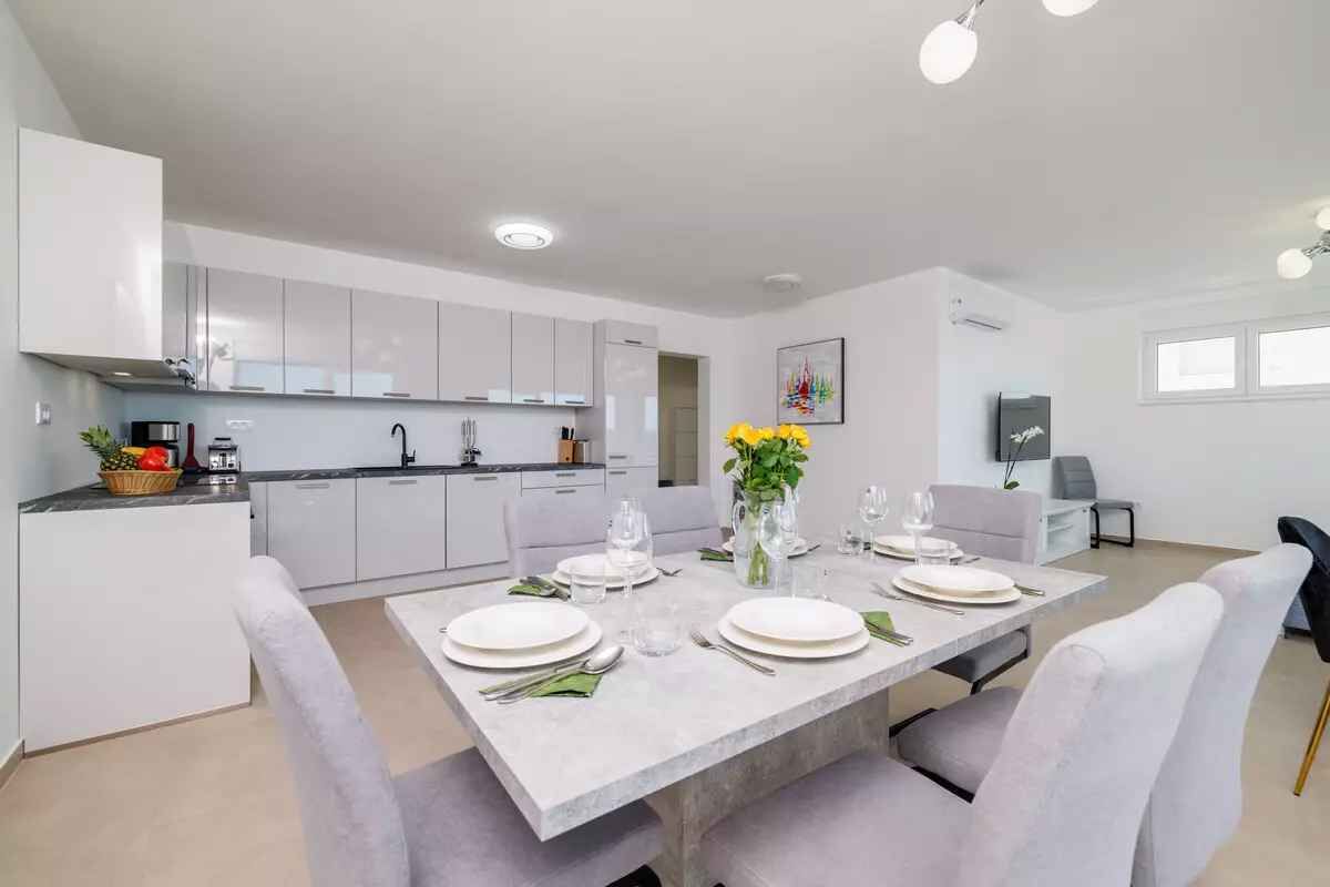 A modern kitchen with white cabinets, a dining table set with plates, glasses, and cutlery, and a vase with flowers.